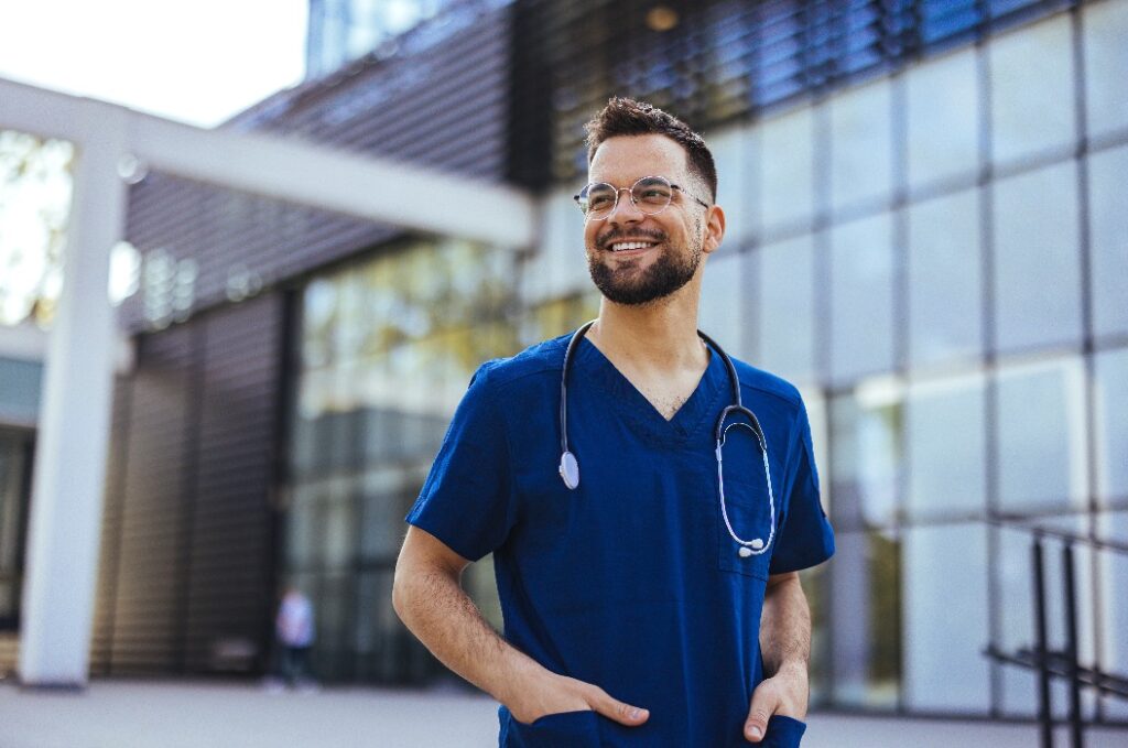 Ein Krankenpfleger steht draußen in OP-Kleidung und mit einem Stethoskop. Er lächelt und schaut in die Kamera. Porträt eines Krankenpflegers. Porträt eines Krankenpflegers mittleren Alters im Krankenhaus